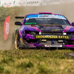 FotoPatrykBierski - Fioletowo-czarny samochód wyścigowy w trakcie driftu, spod kół unosi się kurz, widoczne logo sponsorów i numer 45.