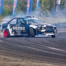 FotoPatrykBierski - Samochód BMW E36 w trakcie driftu na torze, z widocznym dymem z opon, czerwonymi felgami i numerem startowym 179, na tle opon i flag reklamowych.