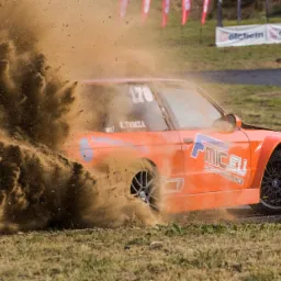 Pomarańczowy samochód wyścigowy w akcji, dynamicznie wzbijający tumany kurzu i ziemi podczas driftu na torze, w tle fotograf w pomarańczowej kamizelce.