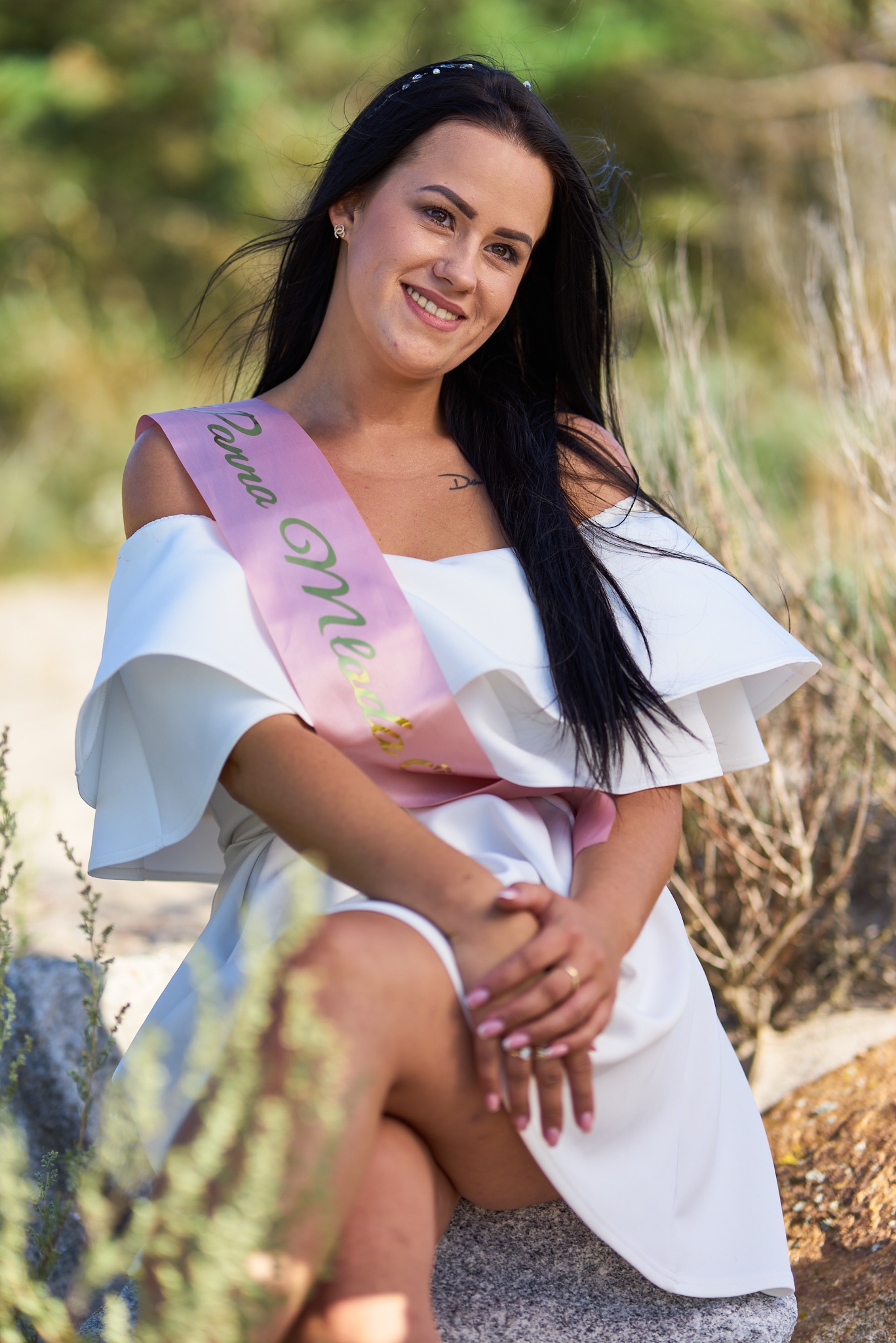 Uśmiechnięta brunetka w białej sukience z odkrytymi ramionami i szarfą z napisem siedzi na kamieniu w plenerze.