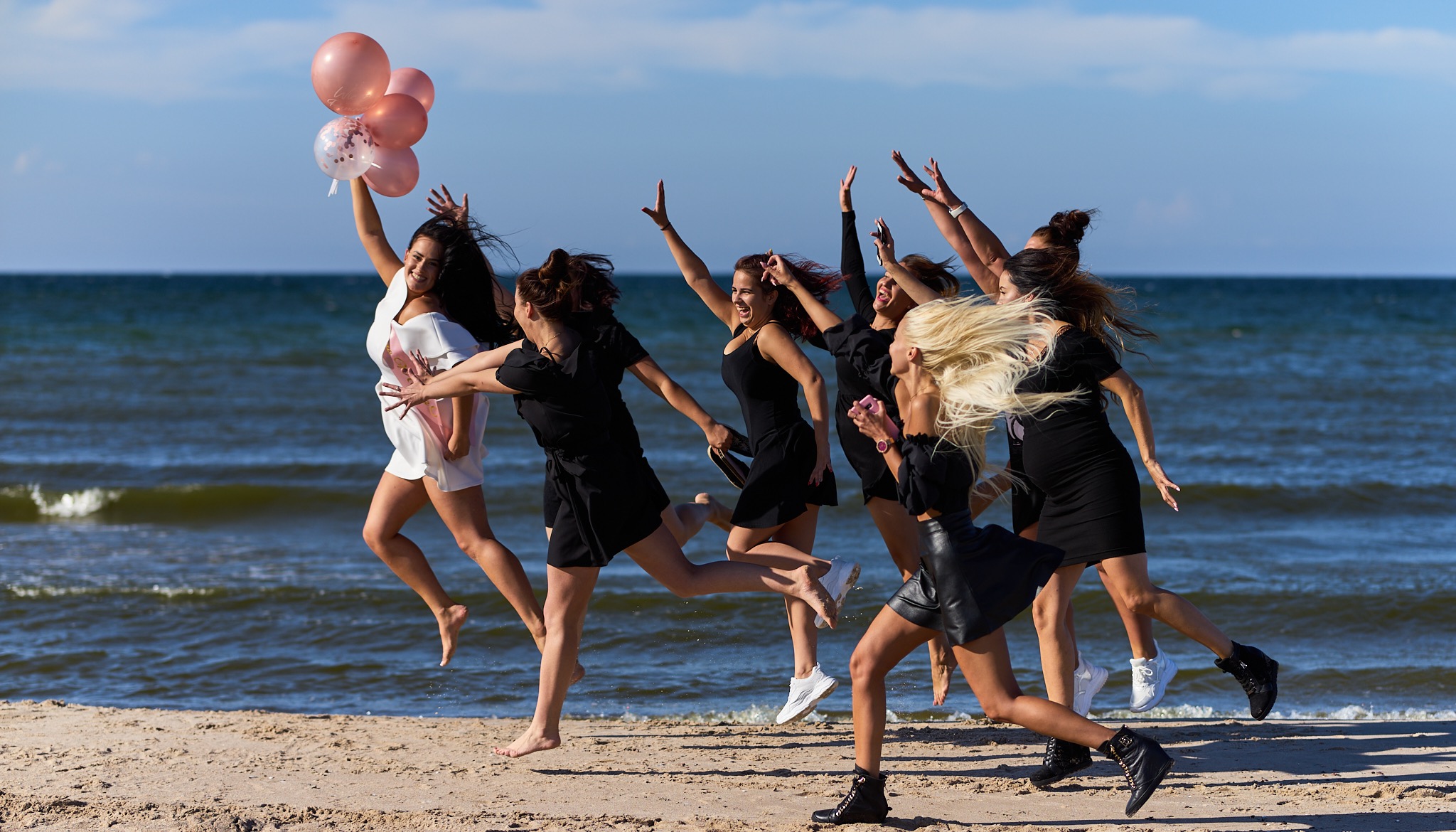 Grupowe zdjęcie z plaży: przyszła mama w białej sukience trzyma balony, a przyjaciółki w czarnych sukienkach radośnie biegną obok, śmiejąc się i wyciągając ręce.