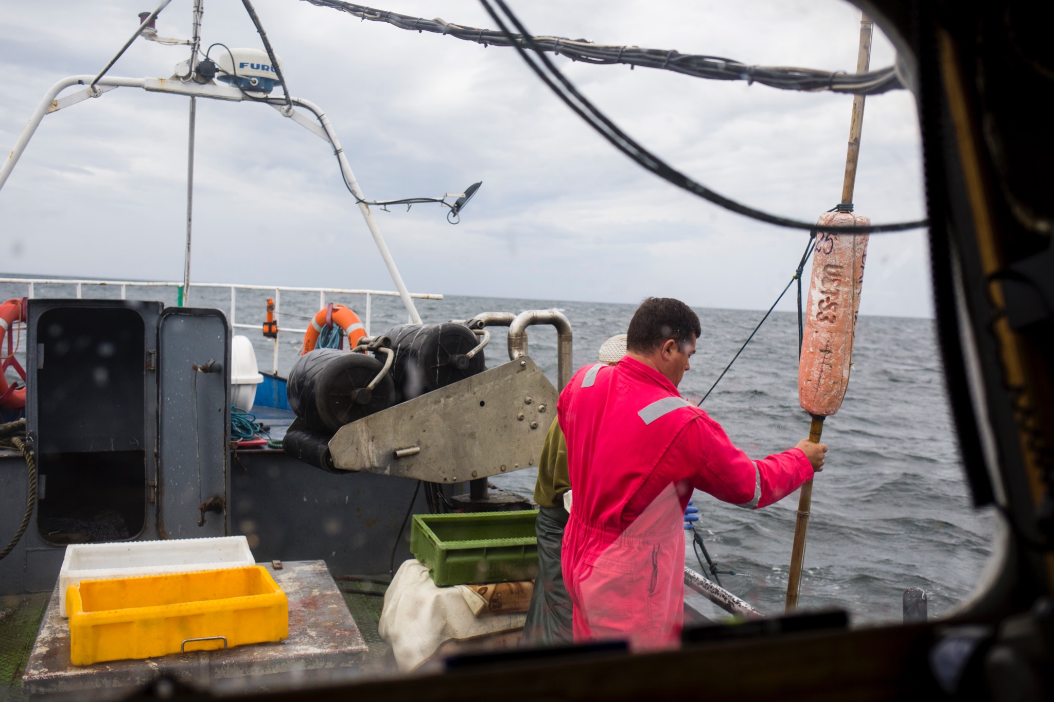 Rybacki kuter na morzu: rybak w czerwonym kombinezonie trzyma boję, widoczne skrzynki na ryby i fragment wyposażenia łodzi.