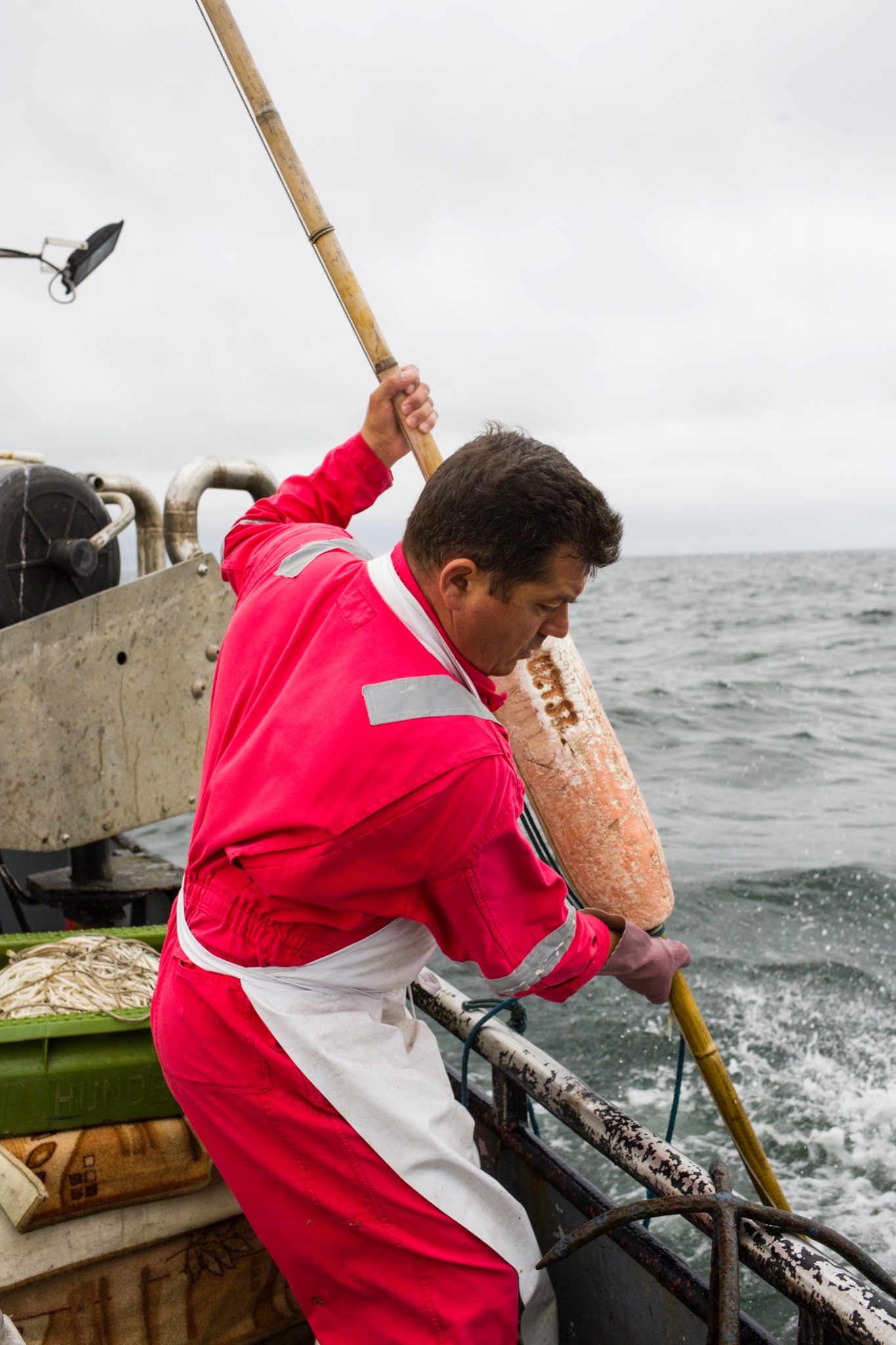Rybolów: Mężczyzna w czerwonym kombinezonie i białym fartuchu, trzymając długi bambusowy kij z pomarańczową boją, pracuje na pokładzie kutra rybackiego na tle wzburzonego morza.
