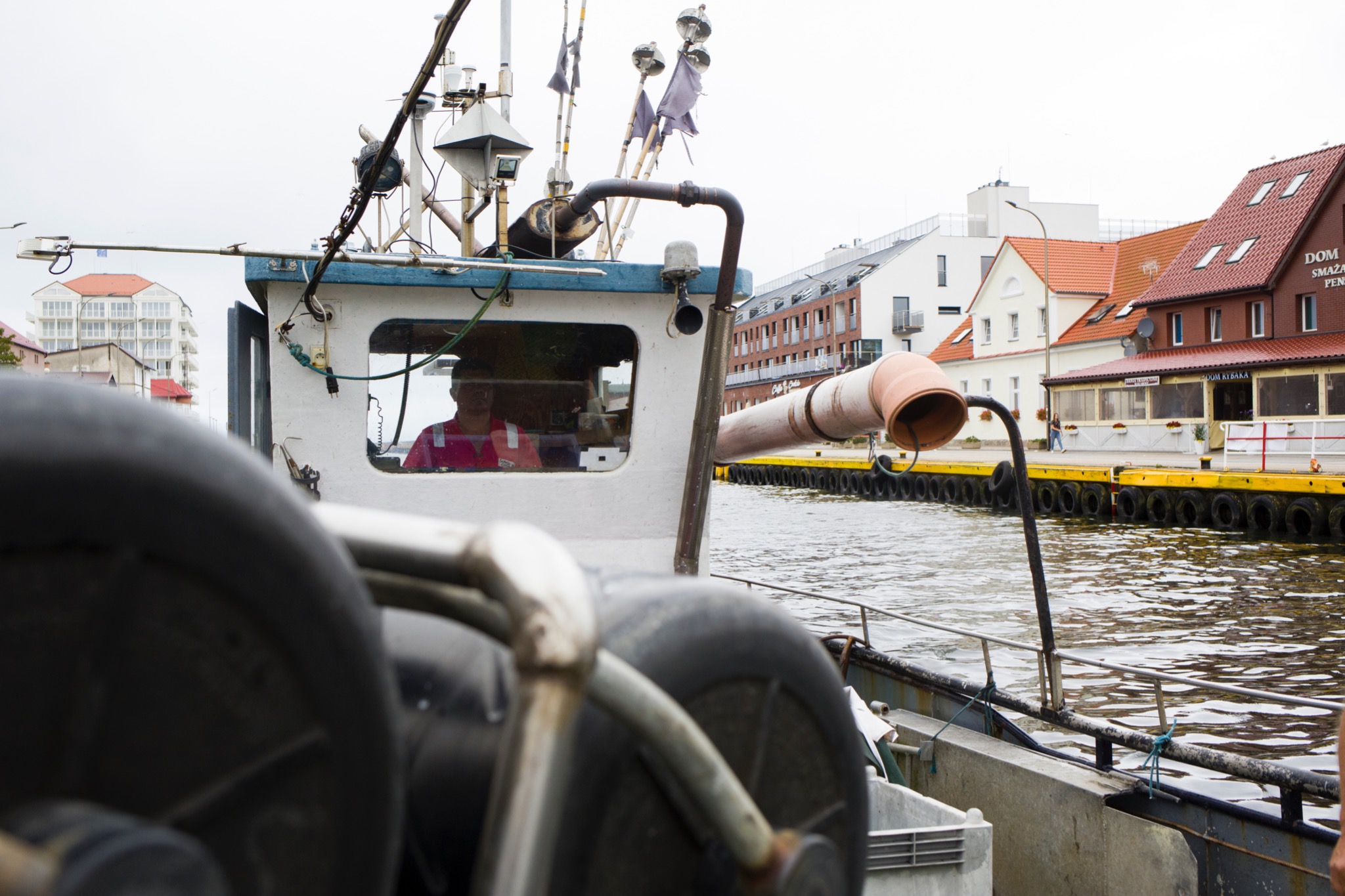 Wnętrze kabiny kutra rybackiego w Koszalinie, widok na nabrzeże z budynkami i pomostem.