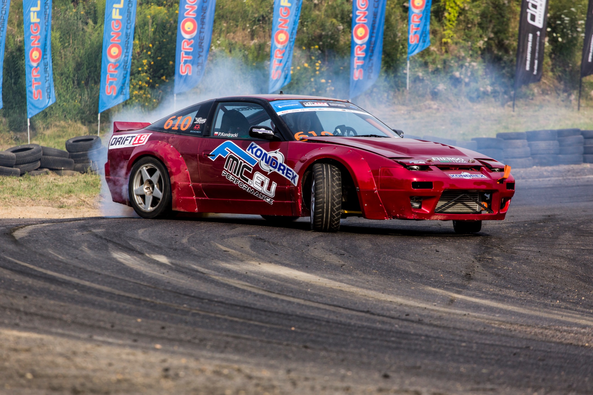 Czerwony samochód do driftu z naklejkami sponsorskimi, w trakcie pokazu, z dymem unoszącym się spod kół, na torze z widocznymi śladami opon.