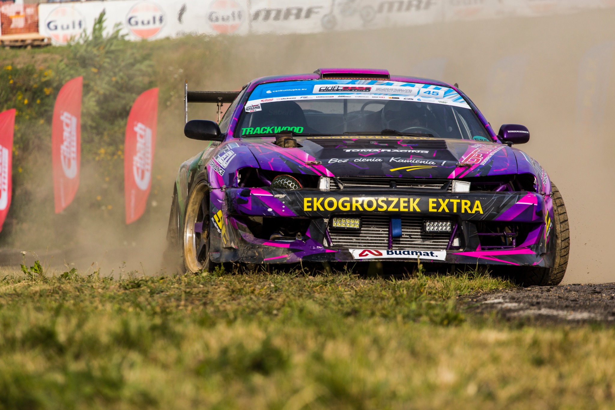 Fioletowo-czarny samochód wyścigowy w trakcie driftu, spod kół unosi się kurz, widoczne logo sponsorów i numer 45.