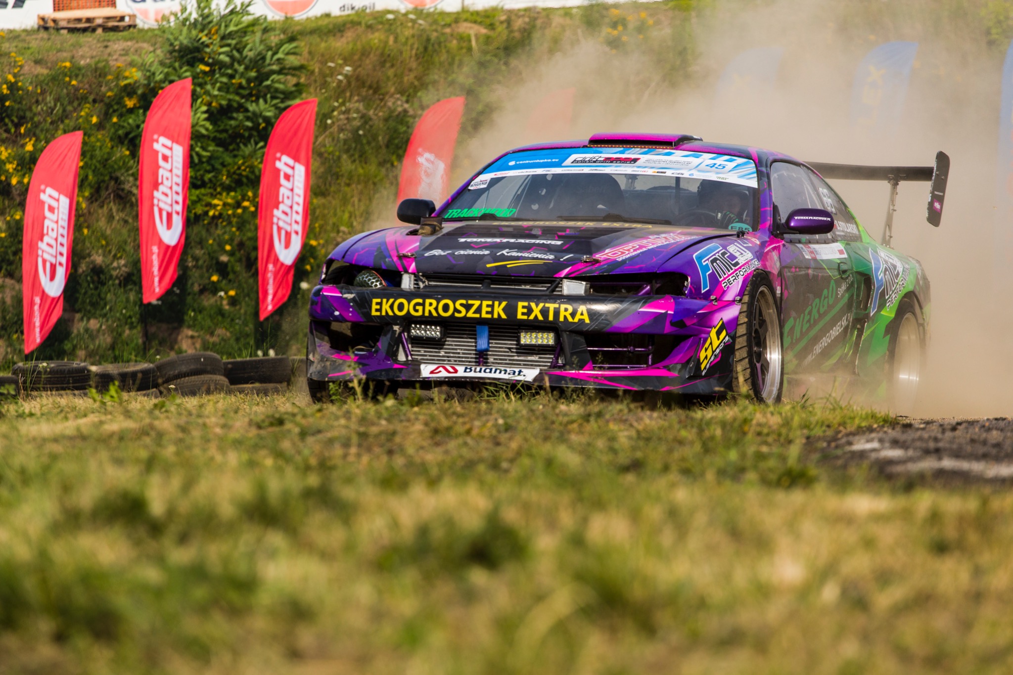 Driftingowy samochód z fioletowo-czarną karoserią w akcji na torze, unoszący tumany kurzu, z widocznymi flagami reklamowymi w tle.