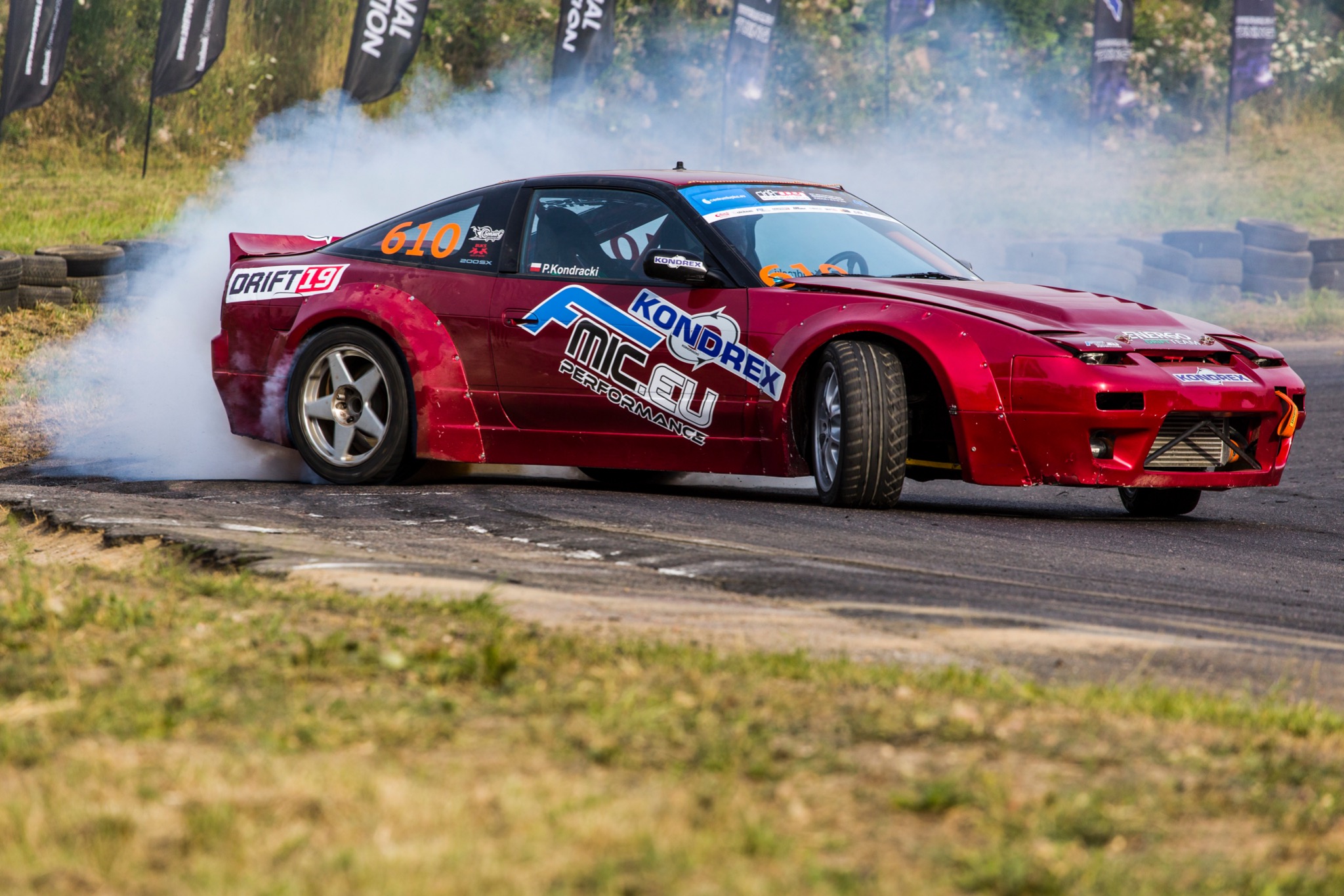 Czerwony samochód do driftu, z numerem 610, bokiem na asfalcie, spod kół unosi się dym, widoczne logo KONDREX i MIC.EU Performance.