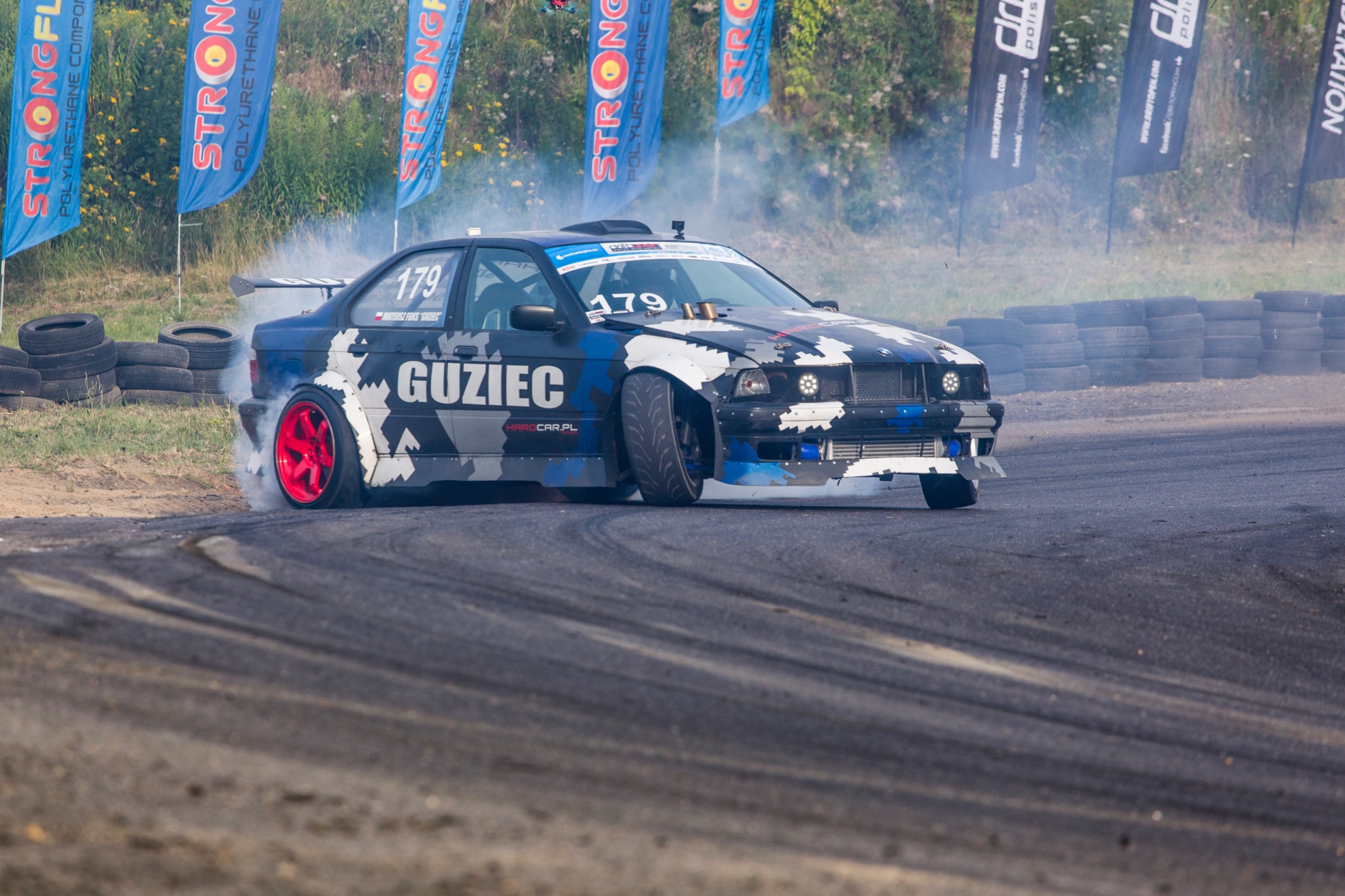 Samochód BMW E36 w trakcie driftu na torze, z widocznym dymem z opon, czerwonymi felgami i numerem startowym 179, na tle opon i flag reklamowych.