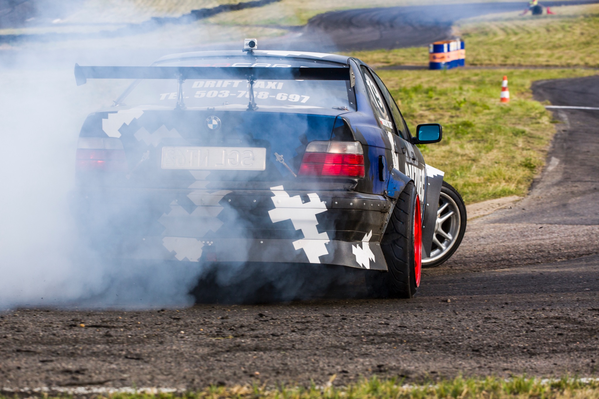 Samochód BMW w kamuflażu driftujący na torze, z tylnego koła unosi się gęsty dym, widoczny spoiler i napis Drift Taxi.