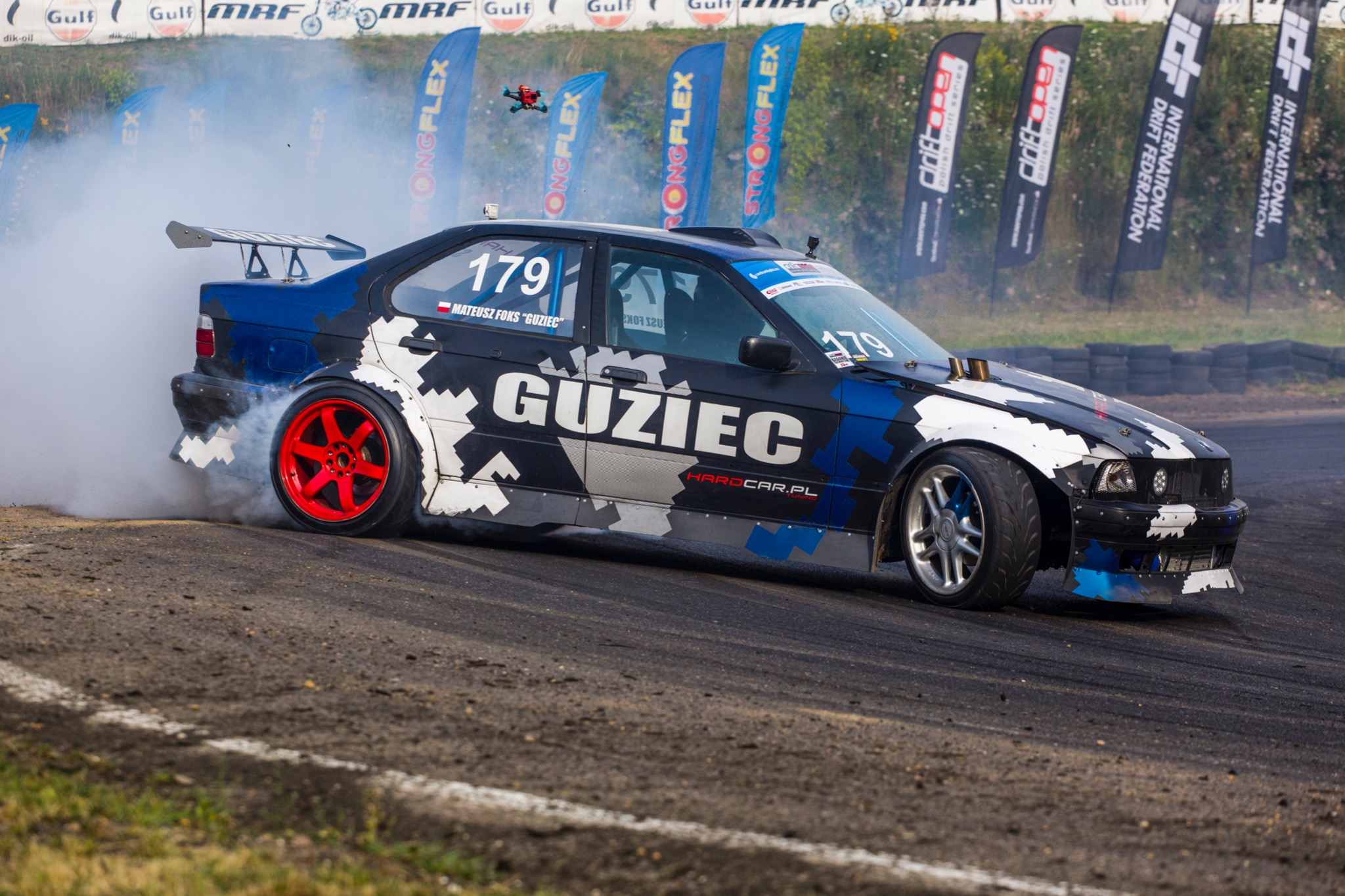 Samochód do driftu w trakcie pokazu, z opon wydobywa się dym, w tle banery reklamowe i opony.