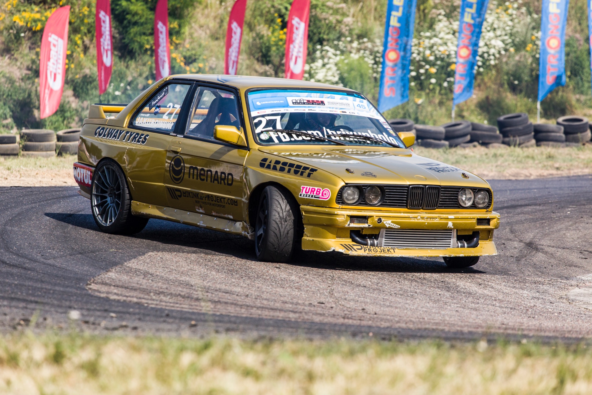 Złoty samochód wyścigowy BMW E30 w poślizgu na torze, z widocznymi oponami, flagami sponsorskimi i naklejkami firm na karoserii.