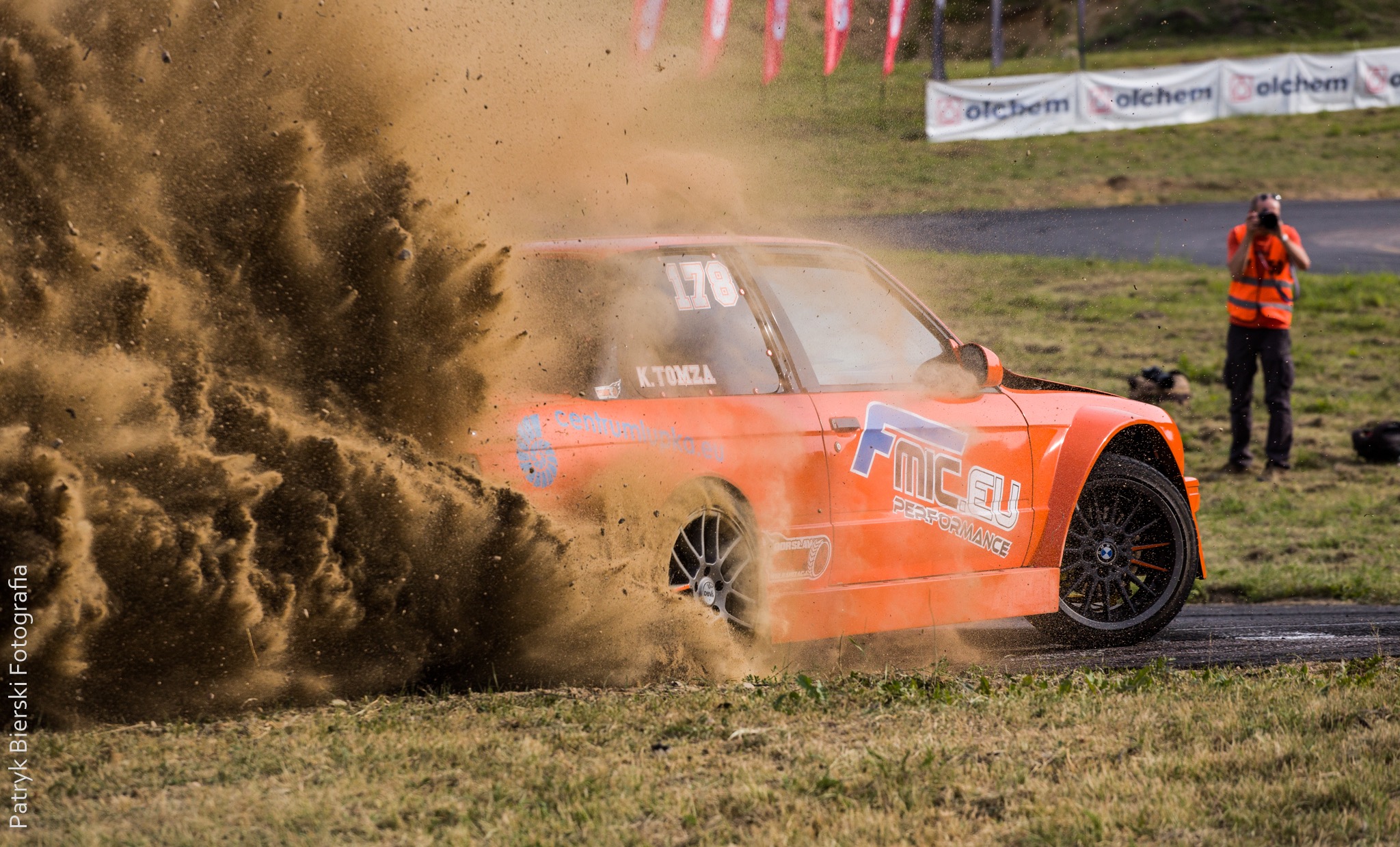 Pomarańczowy samochód wyścigowy w akcji, dynamicznie wzbijający tumany kurzu i ziemi podczas driftu na torze, w tle fotograf w pomarańczowej kamizelce.
