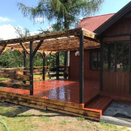 Drewniany taras z zadaszeniem z przezroczystego poliwęglanu, połączony z domkiem, wykończony w brązach i czerniach, z widocznym efektem opalania drewna.