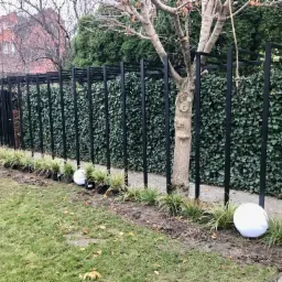 Czarny metalowy stelaż pod pnącza obsadzony bluszczem, białe kule świetlne wzdłuż ścieżki, trawa ozdobna w doniczkach, drzewo z liną.