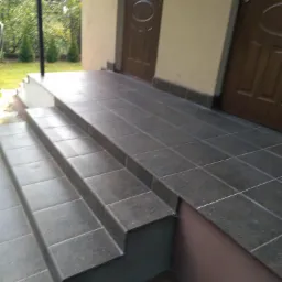 Wykończone płytkami schody wejściowe do domu w Wyszkowie, z widocznymi dwoma drewnianymi drzwiami wejściowymi i fragmentem ogrodu w tle.