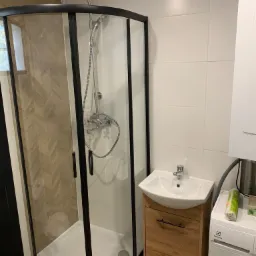 Nowoczesna łazienka z kabiną prysznicową z czarnymi ramami, umywalką na szafce z drewnianym frontem oraz pralką Electrolux z widocznym opakowaniem środków czystości.