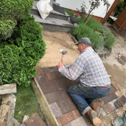 Mężczyzna w kraciastej koszuli i dżinsach, klęczący na ziemi, układa brązową kostkę brukową gumowym młotkiem w ogrodzie obok zielonych krzewów i trawnika.