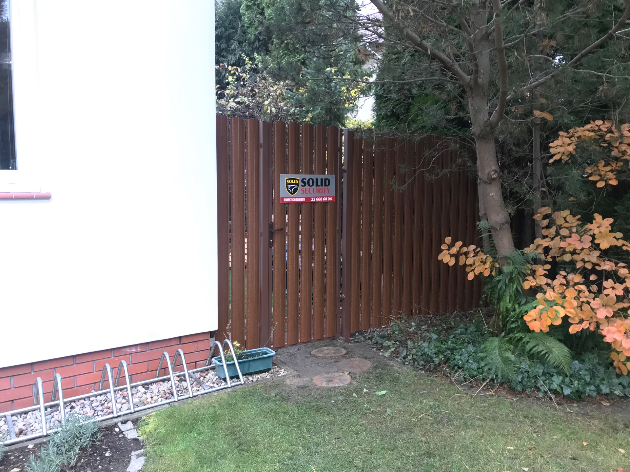 Brązowa furtka z pionowych desek z tabliczką firmy ochroniarskiej Solid Security, umieszczona w ogrodzeniu między budynkiem a drzewami z żółtymi liśćmi.