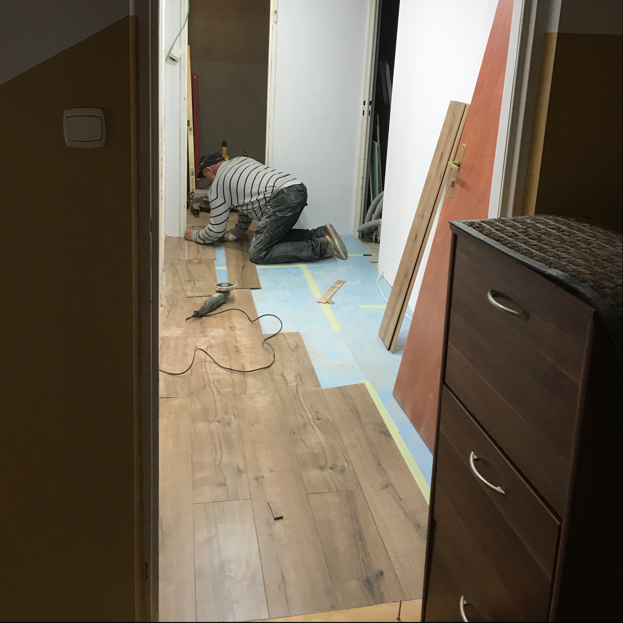 Układanie paneli podłogowych w korytarzu; widoczny fachowiec na kolanach, narzędzia, podkład z taśmą, niedokończona podłoga i fragment szafki z szufladami