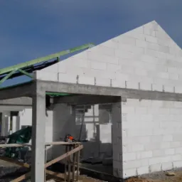 Wznoszenie ścian parterowego budynku z betonu komórkowego, widoczne drewniane elementy konstrukcji dachu, piła łańcuchowa na prowizorycznym stole warsztatowym.