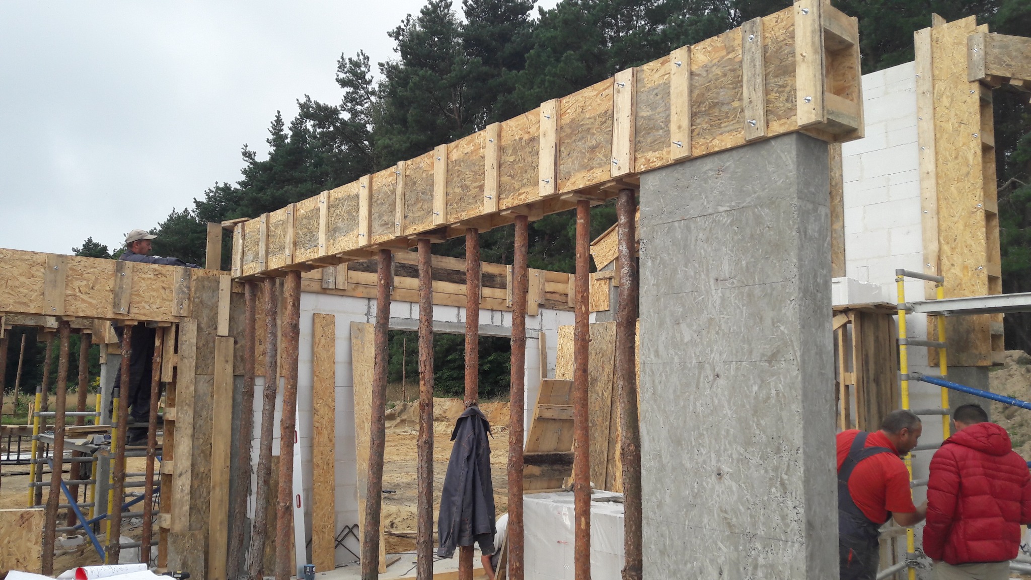 Wznoszenie szkieletu domu z betonu komórkowego i drewnianych szalunków, widoczne pionowe podpory z okrąglaków, pracownicy w ubraniach roboczych, pochmurne niebo w tle.