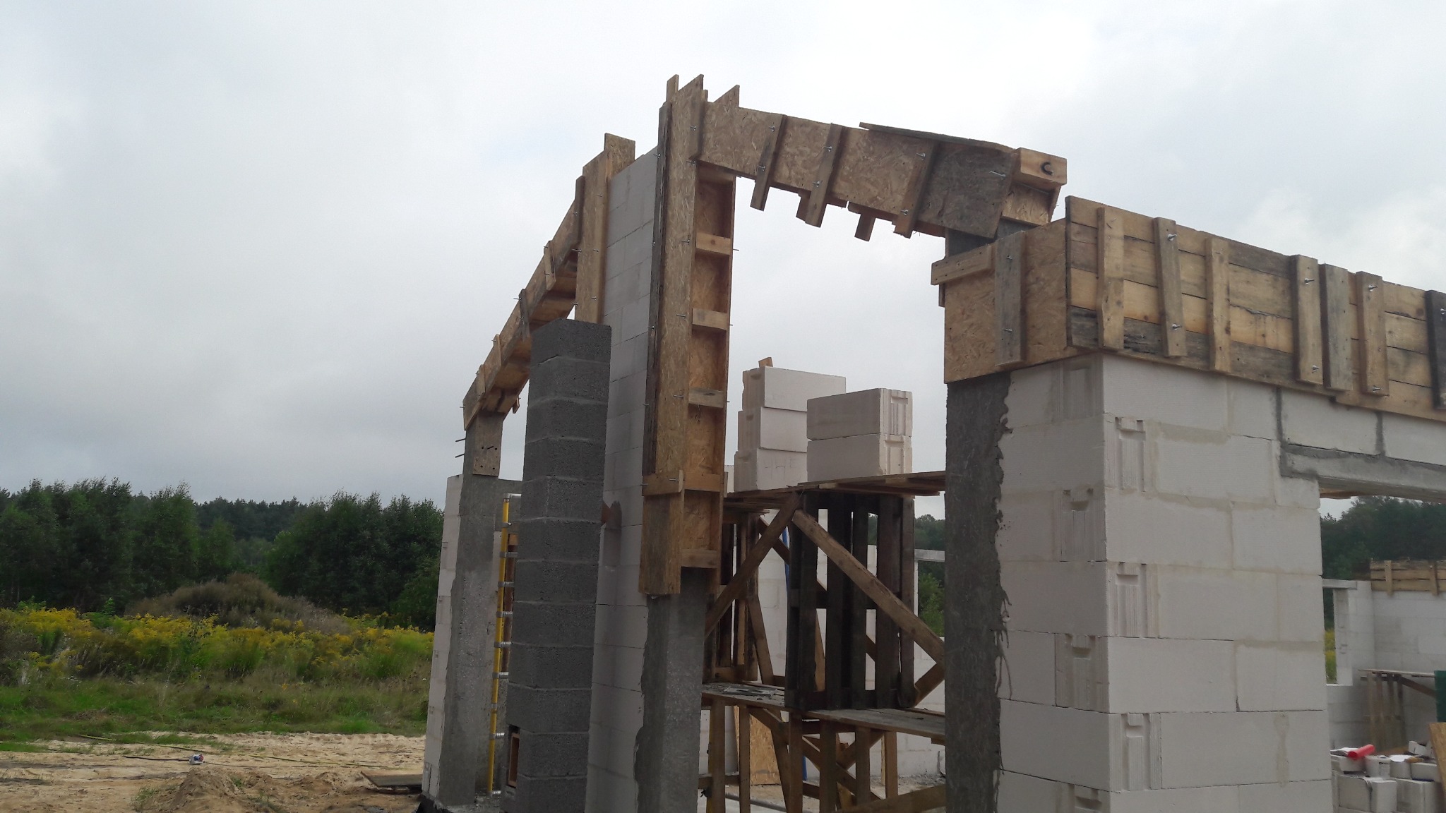 Wznoszenie ścian domu z betonu komórkowego, widoczne szalunki drewniane i rusztowanie, w tle zieleń drzew i pochmurne niebo.