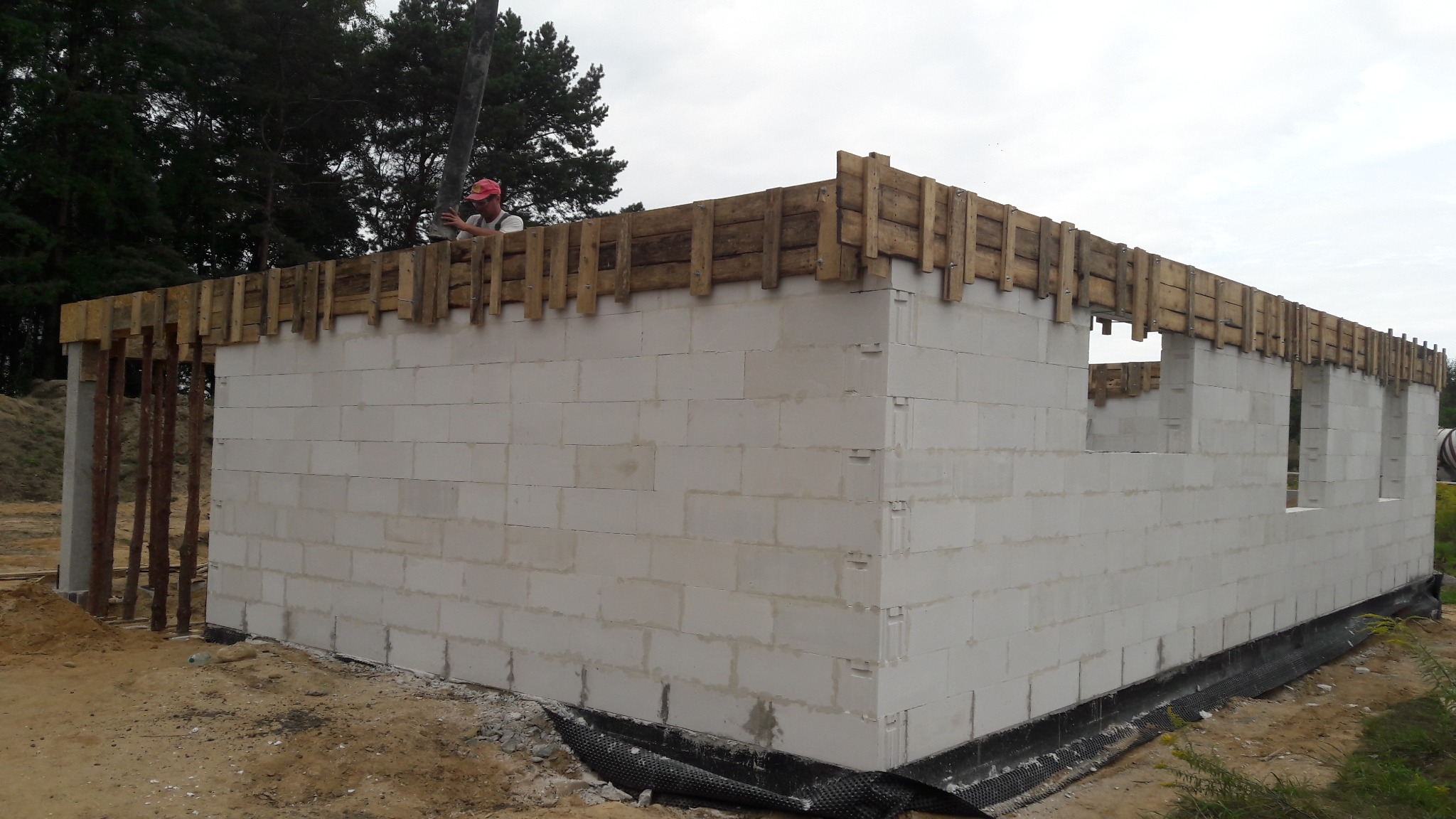 Wznoszenie murów z betonu komórkowego na fundamencie, widoczne szalunki drewniane i pracownik z rurą do wylewania betonu.