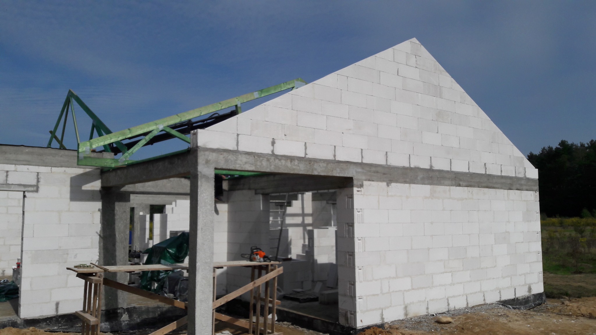 Wznoszenie ścian parterowego budynku z betonu komórkowego, widoczne drewniane elementy konstrukcji dachu, piła łańcuchowa na prowizorycznym stole warsztatowym.