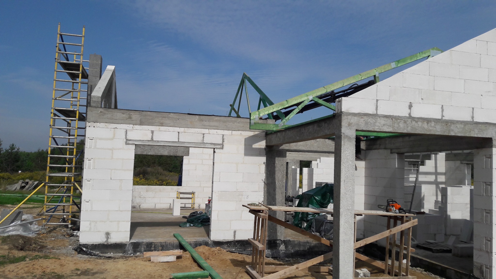 Wznoszenie ścian domu z bloczków betonu komórkowego, widoczne zbrojenia stropu i konstrukcja drewniana dachu, na placu budowy leżą narzędzia i materiały.