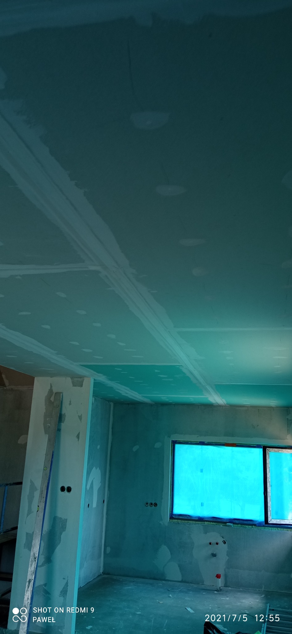 Wnętrze w trakcie prac wykończeniowych: widoczne płyty gipsowo-kartonowe na suficie ze szpachlowanymi łączeniami, ściany po gruntowaniu, okno zabezpieczone folią ochronną, surowa podłoga, widoczna...