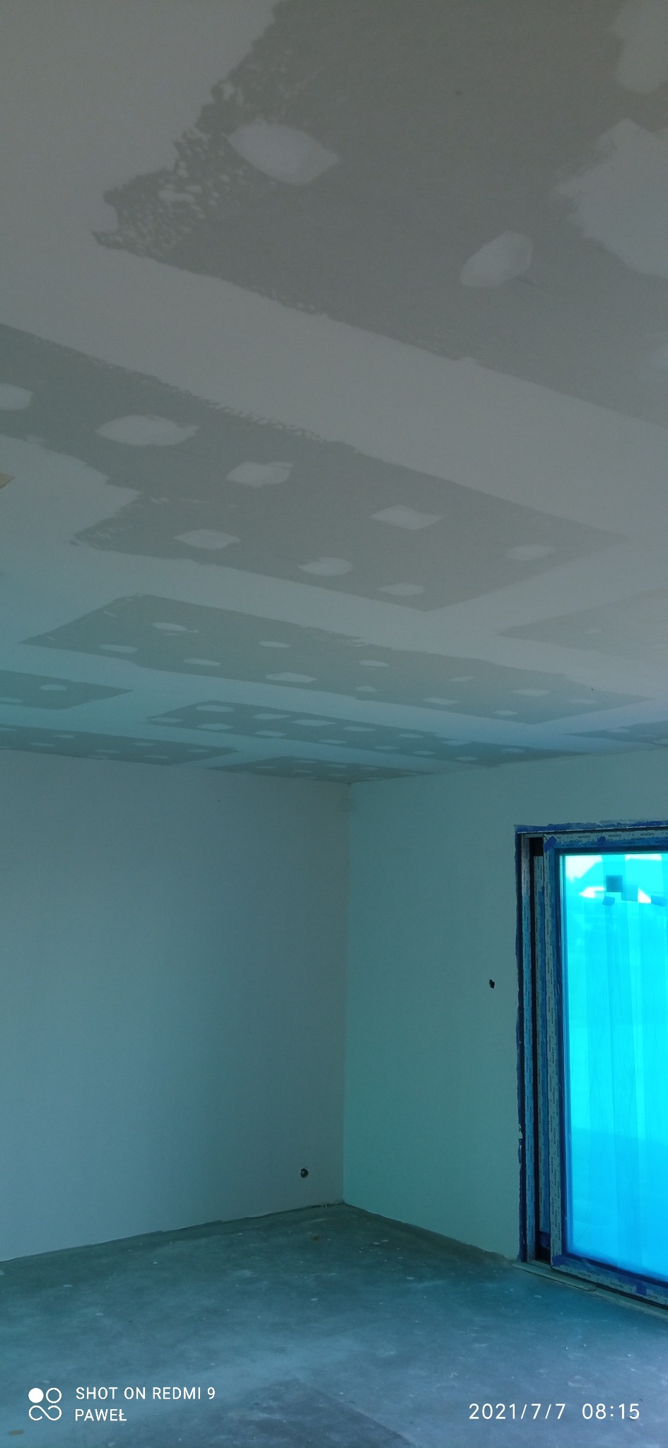 Wnętrze w trakcie szpachlowania sufitu, widoczne nierówności i ślady po szpachli, okno zabezpieczone folią, surowy beton na podłodze.