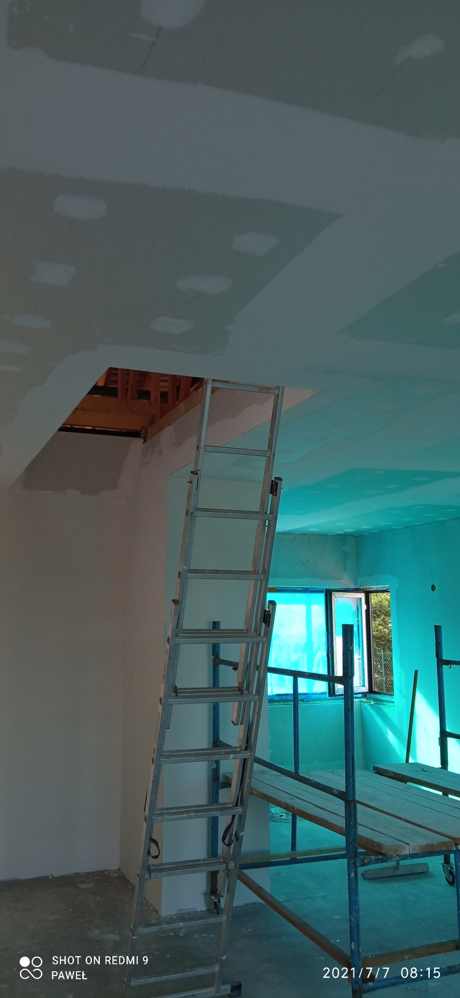 Wnętrze w trakcie prac wykończeniowych: sufit z płyt gipsowo-kartonowych z widocznymi łączeniami i otworami, drabina aluminiowa oparta o ścianę, rusztowanie w tle, okno zasłonięte folią ochronną.