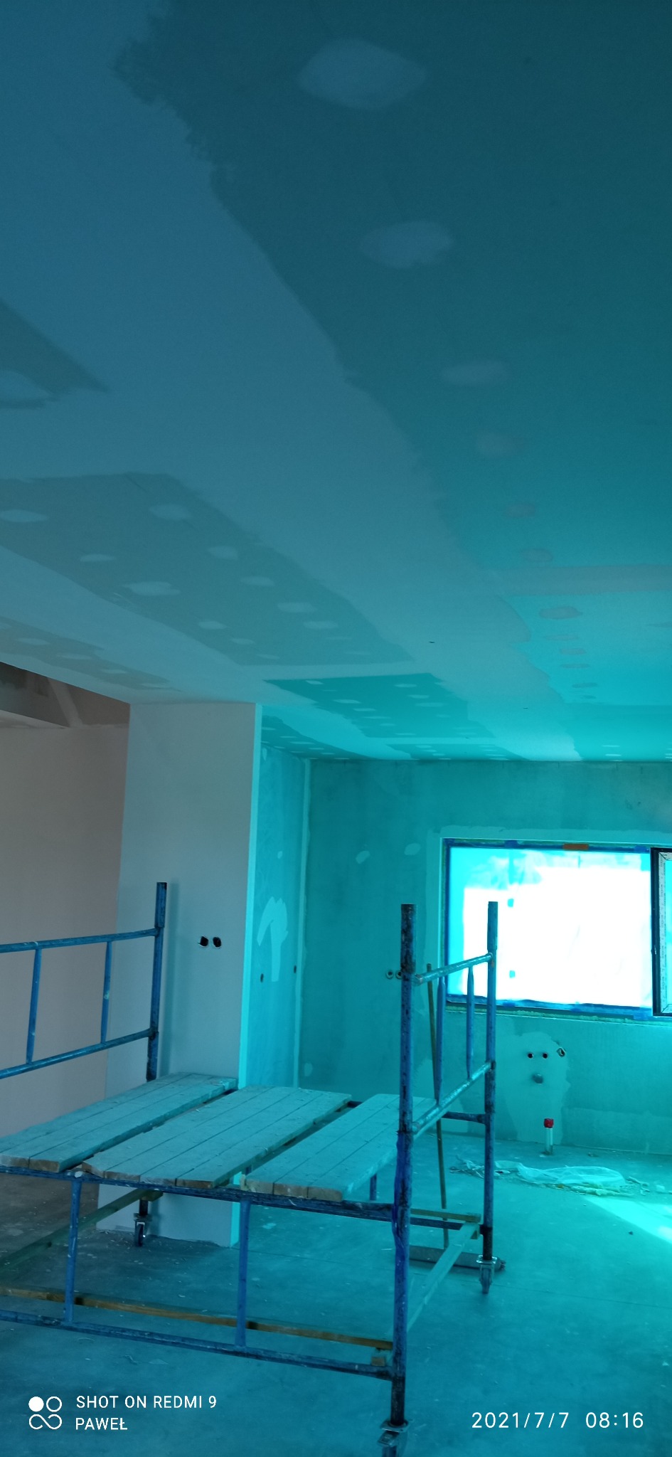 Wnętrze w trakcie prac wykończeniowych: szpachlowany sufit z widocznymi miejscami na oświetlenie, fragment ściany w kolorze brzoskwiniowym, rusztowanie malarskie, okno zabezpieczone folią.