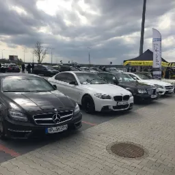 Rząd zaparkowanych, luksusowych samochodów Mercedes i BMW na brukowanym parkingu, z banerami reklamowymi w tle i pochmurnym niebem.