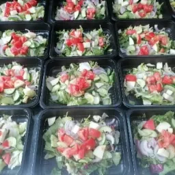 Rząd czarnych pudełek z sałatkami warzywnymi: pomidory, ogórki, cebula, sałata, przygotowanymi do transportu.