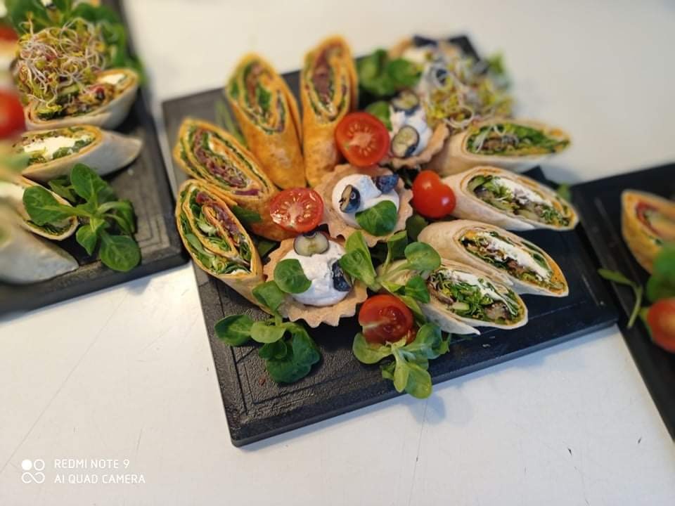 Dekoracyjnie ułożone przekąski cateringowe na ciemnych deskach: tortille z warzywami i serem, mini tartaletki z kremem i borówkami, pomidorki koktajlowe i świeże listki sałaty.