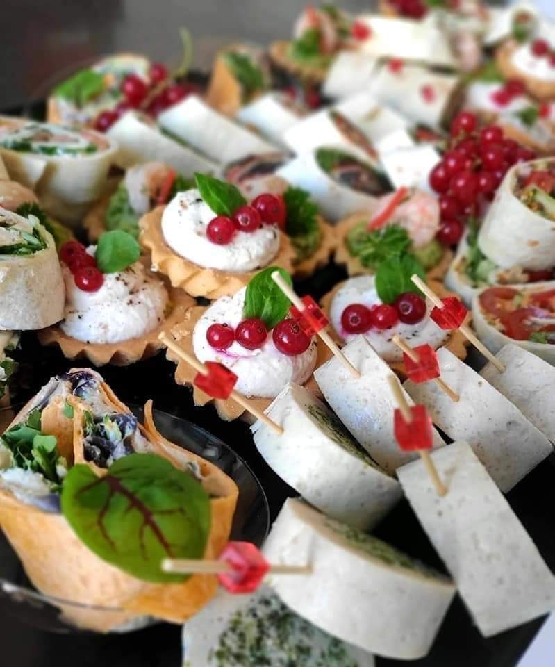 Elegancka kompozycja różnorodnych przekąsek na czarnym talerzu: tartaletki z kremowym serem i czerwoną porzeczką, roladki z tortilli z zielonymi warzywami i trójkątne koreczki z białego sera...