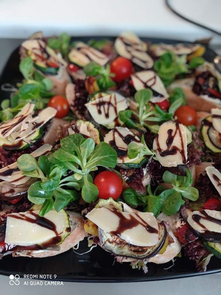 Przekąski na ciemnej tacy: małe kanapki z warzywami (cukinia, bakłażan, pomidorki cherry), serem i sosem balsamicznym, udekorowane roszponką.