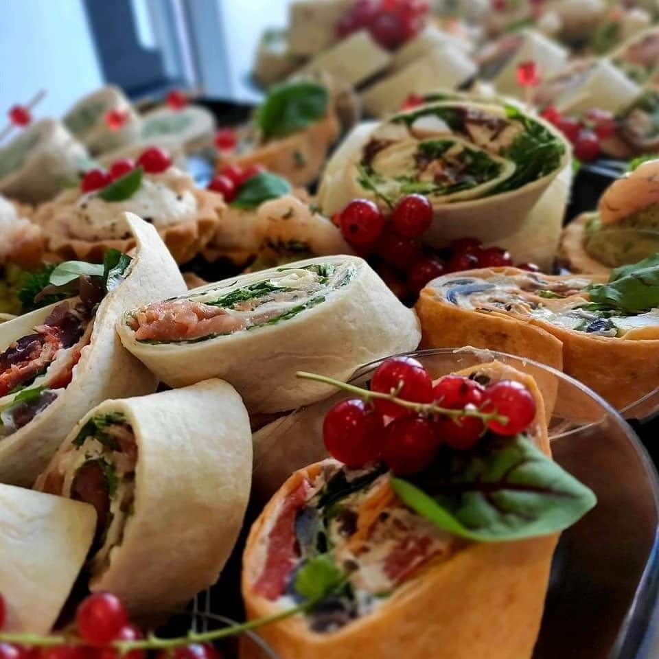 Dekoracyjnie ułożone roladki z tortilli z różnymi nadzieniami, tartaletki z kremem i czerwone porzeczki na czarnej tacy.