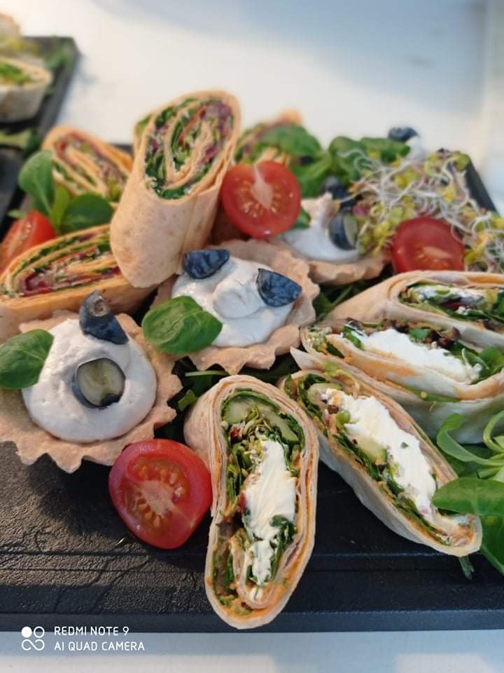 Dekoracyjnie ułożone na czarnym talerzu tortille z różnym nadzieniem, tartaletki z kremem i borówkami, połówki pomidorków koktajlowych i kiełki.