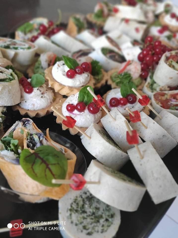 Różnorodne przekąski na catering: tartaletki z kremowym serem i czerwoną porzeczką, roladki z tortilli z różnymi nadzieniami, dekorowane wykałaczkami z czerwonymi elementami.
