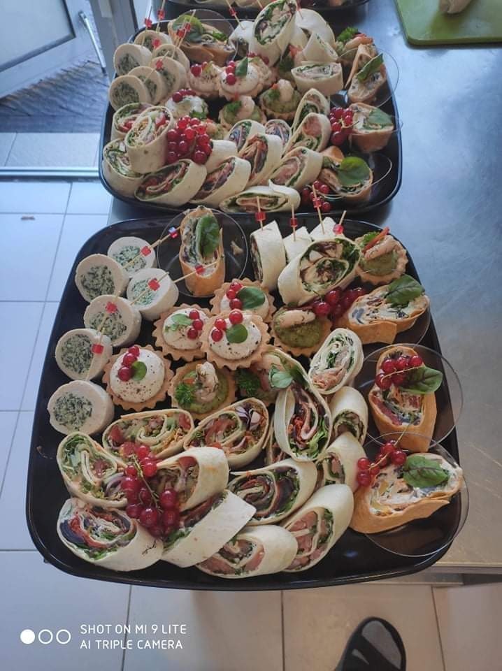 Dwie czarne tace z różnorodnymi przekąskami cateringowymi: roladki z tortilli z widocznym wnętrzem z warzywami i mięsem, mini tartaletki z kremowym nadzieniem i dekoracją z czerwonych porzeczek...