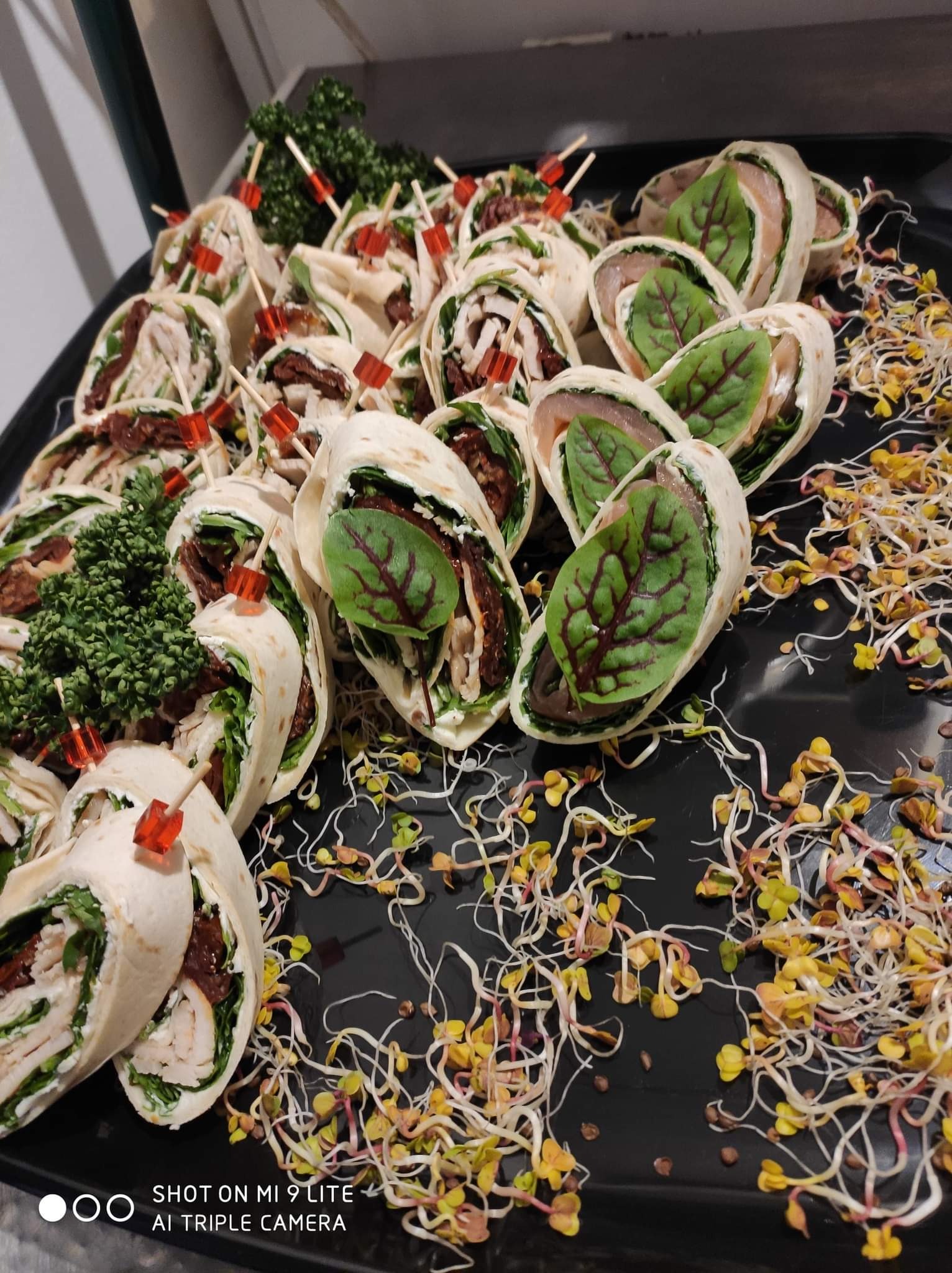 Czarny talerz z roladkami z tortilli z różnymi nadzieniami: kurczak z suszonymi pomidorami i łosoś z zielonymi liśćmi, udekorowane kiełkami i gałązkami zieleni, zabezpieczone czerwonymi wykałaczkami.