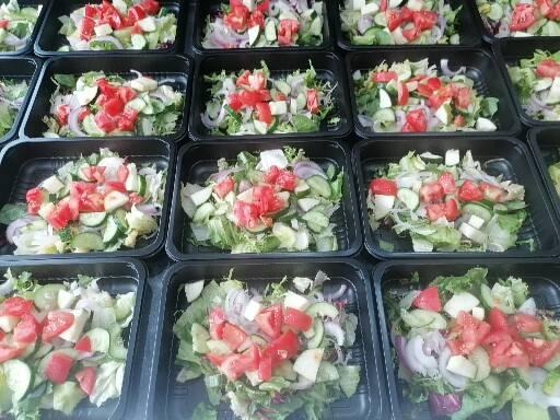 Rząd czarnych pudełek z sałatkami warzywnymi: pomidory, ogórki, cebula, sałata, przygotowanymi do transportu.