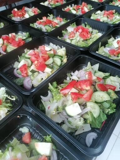 Porcje sałatek warzywnych z pomidorami, ogórkami i czerwoną cebulą w czarnych, plastikowych pojemnikach, ułożone rzędami.