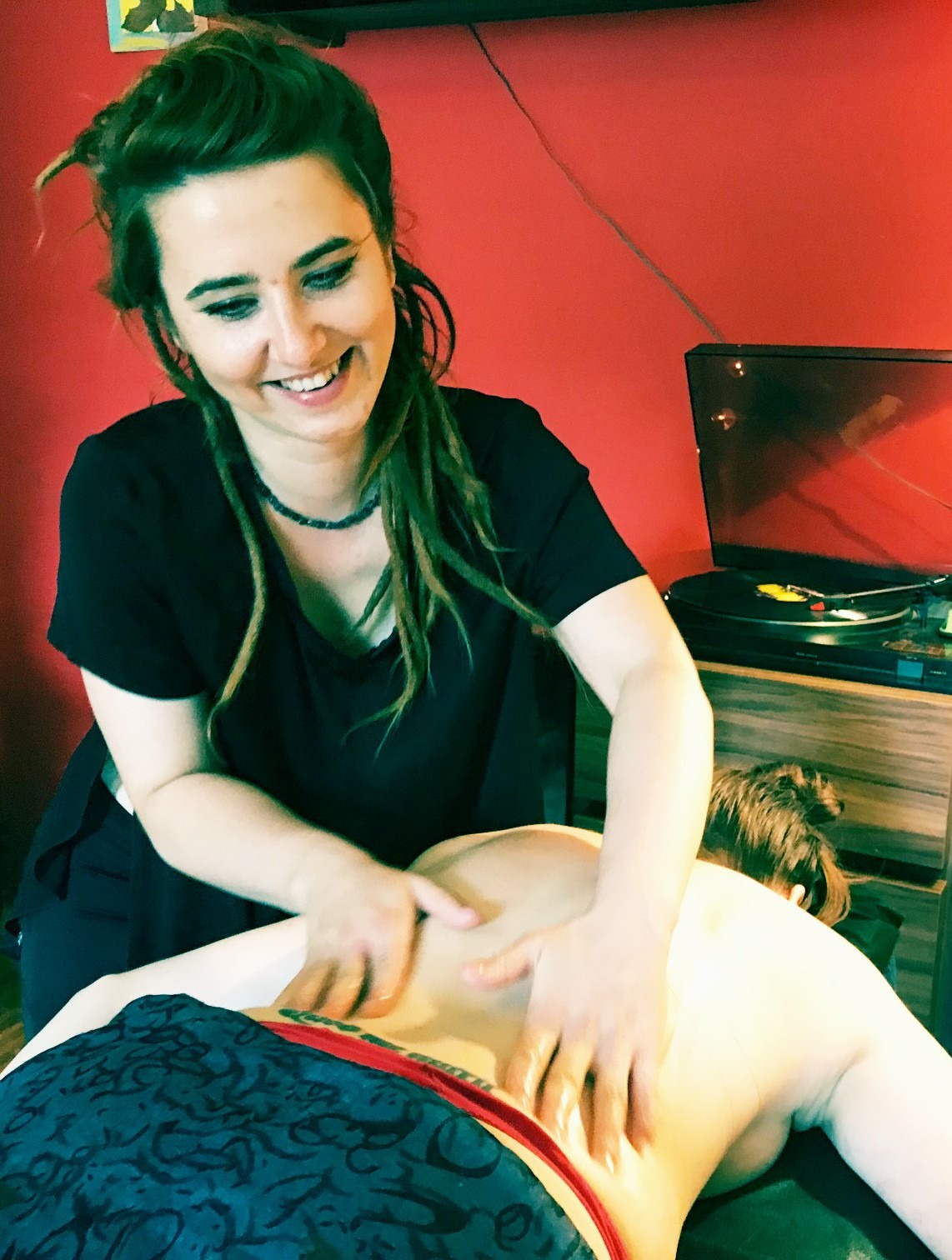 Uśmiechnięta masażystka z dredami wykonuje masaż pleców pacjentki na tle czerwonej ściany i gramofonu.