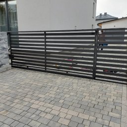 MAZO Creator - Nowoczesna, przesuwna brama wjazdowa z poziomych, grafitowych profili stalowych, widok z zewnątrz na kostkę brukową i fragment budynku.