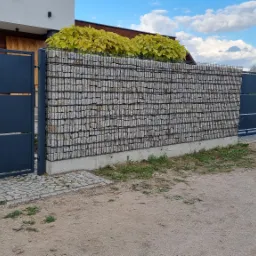 Nowoczesna brama wjazdowa i furtka z metalowymi panelami w kolorze grafitowym, osadzone w gabionowych słupkach wypełnionych kamieniami, z widocznym fragmentem domu w tle.