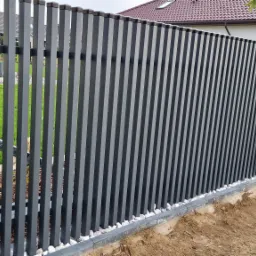 Nowoczesne ogrodzenie panelowe z metalowymi sztachetami w kolorze grafitowym, osadzone na słupkach z cegły klinkierowej, z drobnym białym grysem u podstawy i widocznym trawnikiem w tle.